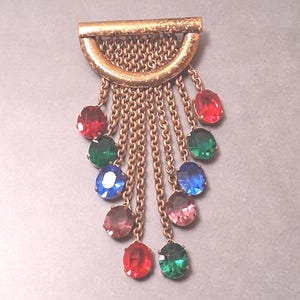 May include: A gold-toned metal brooch featuring a semi-circular top bar with a textured finish. Attached to the bar are multiple cascading gold-toned chains of varying lengths, each ending in a faceted oval gemstone in red, green, blue, or purple.