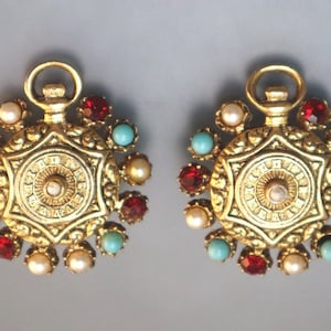 May include: A pair of gold-tone earrings with a circular design. Each earring features a central star-shaped pendant with a smaller circle in the center. The pendant is surrounded by a ring of smaller circles, each with a red, turquoise, or white stone. The earrings are suspended from a loop.