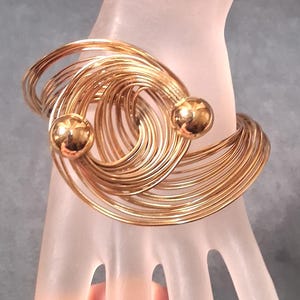 May include: A gold-toned cuff bracelet constructed from multiple thin, overlapping wire loops that form a wide, circular band. The design features two polished, spherical gold-toned beads positioned at the open ends of the wire structure.