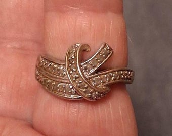 May include: Silver-toned ring with a knot design, encrusted with small, clear gemstones. The ring's band and knot are detailed with the gemstones, creating a sparkling effect. The ring is shown on a finger.