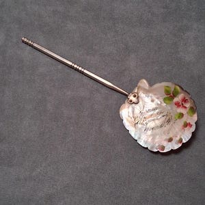 May include: A decorative silver-toned seashell-shaped spoon with a long handle. The shell is adorned with painted pink and green flowers and handwritten text. The spoon rests on a gray surface.