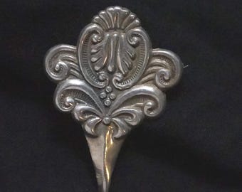 May include: Silver brooch with an elaborate, symmetrical design. The brooch has a central floral motif with detailed scrollwork and a pointed, teardrop-shaped base. The design is highly detailed and appears to be vintage.