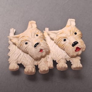 May include: A vintage brooch featuring two cream-colored Scottish Terrier dogs. The dogs have black eyes and noses, with red mouths. The brooch is made of a textured material and is set against a gray background.