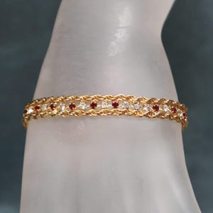 May include: Gold-toned bracelet with a braided design, adorned with alternating red and clear gemstones. The bracelet is displayed on a translucent surface, showcasing its intricate details and elegant style. A classic piece of jewelry.