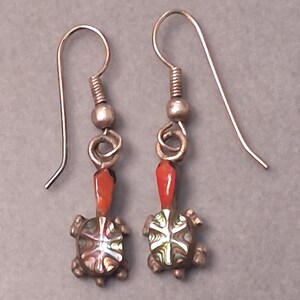 May include: A pair of drop earrings featuring turtle-shaped charms with iridescent, multi-colored shells and bright orange heads. The earrings are suspended from silver-toned metal hooks with a small bead accent above each turtle figure.