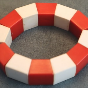 Könnte beinhalten: Ein rotes und weißes Plastikarmband aus ineinandergreifenden Teilen. Das Armband hat die Form eines Kreises und ist an einem Ende offen.