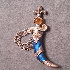 May include: A decorative dagger pendant with a silver-tone finish, featuring a blue enamel handle and adorned with clear and amber-colored crystals. The pendant includes a chain for wearing.