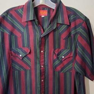 May include: A short-sleeved button-down shirt with a vertical striped pattern in burgundy, navy, and green. The shirt has two chest pockets with button closures and a pointed collar. The brand label is visible on the collar.
