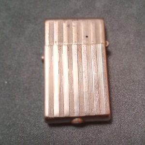 May include: A vintage silver-toned cigarette case with a rectangular shape. The case features vertical, textured lines across its surface. It has a small hinge and a clasp on the bottom. The case is likely made of metal.