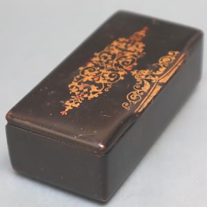 Puede incluir: Una caja rectangular de color marrón oscuro con tapa abatible. La superficie superior presenta un intrincado patrón de filigrana alargado de color dorado con pequeños acentos rojos. La caja tiene una construcción sólida y lisa.
