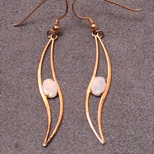 May include: A pair of gold-colored earrings with a curved, open design. Each earring features a small, oval-shaped, off-white stone set in the center. The earrings have a hook closure.