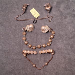 Könnte beinhalten: Drei Vintage-Schmuckstücke: ein Paar silberfarbene Kragenstäbe mit Kette, ein Paar Ohrringe mit Perlen und Strasssteinen und eine Brosche mit Perlen und Strasssteinen mit Kette. Der Schmuck wird auf einer grauen Oberfläche präsentiert.