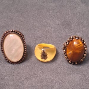 May include: Three rings are displayed on a gray surface. The first ring has an oval, off-white stone set in a bronze-colored metal frame. The second ring is yellow with a dark brown design. The third ring has a brown stone with a bronze-colored frame.