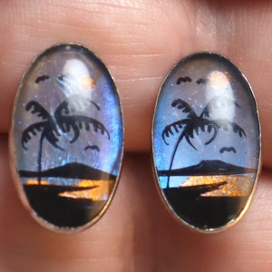 May include: A pair of oval-shaped earrings with a blue and purple iridescent background. The earrings feature a silhouette of palm trees and a sunset on a beach.
