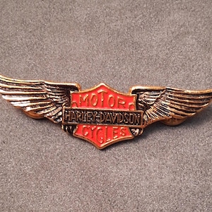 May include: A metallic pin featuring a central shield with the text MOTOR HARLEY-DAVIDSON CYCLES in black and red enamel. The shield is flanked by two detailed, textured bronze-colored wings extending outward on both sides of the emblem.