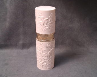May include: A cylindrical, pale pink perfume bottle with embossed floral and bird designs. A gold band encircles the middle, with the words "L'Air du Temps" in gold lettering. The bottle has a rounded top.