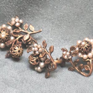 Peut inclure: Une paire de boucles d'oreilles de style vintage avec un motif floral. Les boucles d'oreilles ont une base en métal de couleur bronze avec des branches et des feuilles délicates. Chaque boucle d'oreille a une forme de fleur centrale entourée de petites perles blanches.