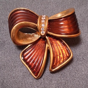 Könnte beinhalten: Vintage Brosche in Schleifenform mit goldfarbenem Rahmen und rotbrauner Emaille-Oberfläche. Die Mitte der Schleife ist mit einer Reihe funkelnder, klarer Strasssteine verziert, die diesem klassischen Accessoire Eleganz verleihen.