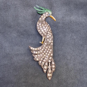 May include: A silver-toned bird brooch with a green crest, blue eye, and yellow beak. The bird's body and tail are encrusted with clear, sparkling crystals. The brooch is set against a gray background.