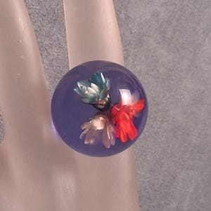 May include: A clear, round ring with a purple hue, encasing a cluster of colorful dried flowers. The flowers include red, white, and teal blooms, creating a unique and artistic accessory. The ring is a statement piece.