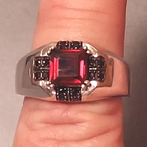 Könnte beinhalten: Ein silberner Ring mit einem großen, rechteckigen, roten Edelstein in der Mitte, umgeben von kleineren schwarzen Steinen. Der Ring hat ein breites Band und eine polierte Oberfläche, die ein klassisches Design zeigt.