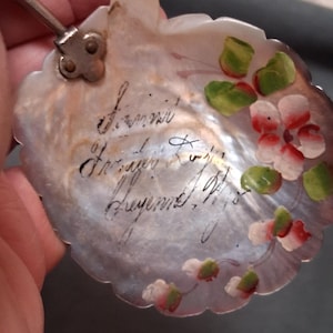 May include: A mother-of-pearl spoon with a scalloped edge and a floral design. The spoon features hand-painted pink and white flowers with green leaves. The handle is attached with a silver-colored metal piece. The spoon has handwritten text.