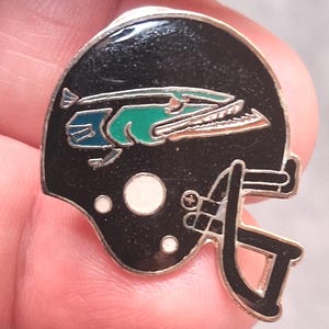 May include: A black enamel pin shaped like a football helmet. The helmet features a stylized green and blue fish graphic on the side, a black face mask, and three small white circular accents near the bottom edge, all outlined in silver metal.