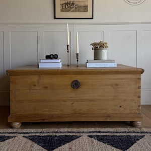 May include: A rectangular light brown wooden storage chest with a central circular metal keyhole plate and four small bun feet. The top surface holds two white taper candles in brass holders, two stacked books, and a small ceramic pot with dried flowers.