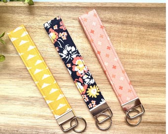 May include: Three fabric key fobs with metal hardware. One is yellow with white cloud shapes, another is navy with floral patterns, and the third is peach with small flower shapes. The key fobs are displayed on a wooden surface.