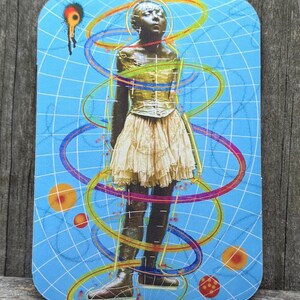 May include: A rectangular sticker featuring a bronze-colored statue of a person in a gold top and cream skirt, surrounded by colorful swirling lines and orbs on a blue grid background. A black and red shape is in the upper left corner.