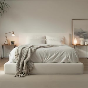 Pode incluir: Uma configuração moderna de quarto com uma cama branca, cabeceira e base a condizer. Uma manta de malha cinza claro é drapeada sobre a cama. Duas pequenas mesas de cabeceira de madeira com lâmpadas e decoração ladeiam a cama, e uma pintura de paisagem emoldurada está pendurada na parede.