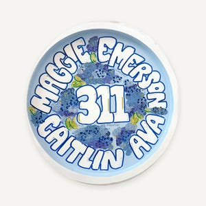 May include: A round, light blue decorative tray featuring the text MAGGIE EMERSON, CAITLIN AVA, and 311 in bold white lettering. The design includes illustrations of blue hydrangea flowers and small green leaves surrounding the central number.