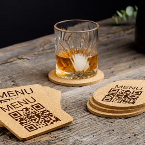 May include: A glass of amber liquid with ice rests on a round cork coaster. Several cork coasters with the word "MENU" and a QR code are stacked on a wooden surface. The coasters are light brown.