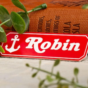 May include: Red and white sign with a white anchor and the text "Robin" in white cursive lettering.