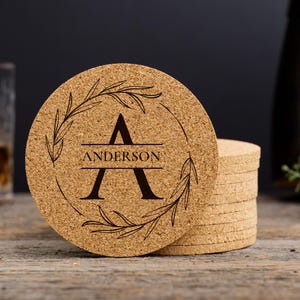 May include: Circular cork coasters with a personalised design. The coasters display a large letter "A" with the name "Anderson" beneath, encircled by a decorative leaf wreath. A stack of coasters is visible.