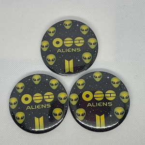Peut inclure: Trois boutons ronds noirs avec un motif de visage d'extraterrestre jaune et le mot "ALIENS" en jaune. Les boutons ont un fond étoilé et un motif de rayures verticales jaunes.