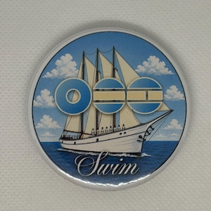 May include: A round, silver-edged button with a nautical theme. The design features a white sailboat on blue water under a blue sky with white clouds. The word "Swim" is written in cursive below the boat.