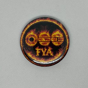 May include: Round button with a fiery design. The button features a black background with three stylized symbols and the letters "FYA" in a flame-like font. The outer edge of the button has a ring of orange and yellow flames.