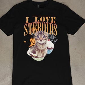 Pode incluir: Camiseta preta com uma estampa gráfica de um gato sentado em uma xícara com um cupcake. O texto "I LOVE STEROIDS" está impresso na camiseta.