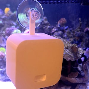 May include: A white, square-shaped device with a suction cup mount attached to a glass aquarium. The device has a black cord extending from the bottom.