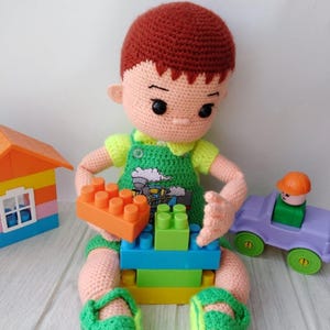 May include: A handmade crochet doll of a child wearing green overalls and sandals, holding orange and green building blocks. The doll has red hair and is sitting next to a toy car and a toy house.