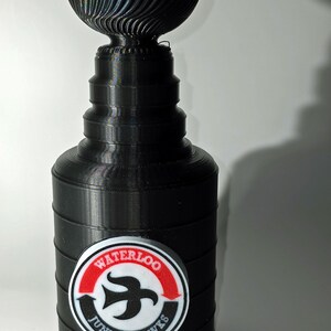 May include: A black plastic replica of the Stanley Cup trophy with a red, white, and black logo that says "Waterloo Junior Hawks".