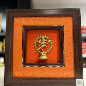 Op de afbeelding: Een ingelijst kunstwerk met een goudkleurig beeld van een dansende figuur, tegen een levendige oranje achtergrond met een patroon. Het beeld is omlijst in een donkerbruine houten lijst, wat een opvallend contrast creëert. Het kunstwerk is vierkant.