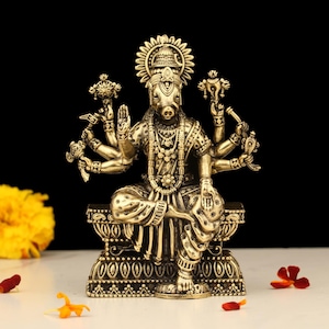 May include: A detailed brass statue of a deity with a boar's head and multiple arms, seated on an ornate base. The statue is adorned with intricate jewelry and a halo, set against a black background. Decorative elements include flowers.