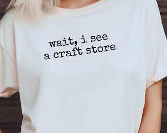 Può includere: T-shirt color crema con la scritta nera "wait, i see a craft store". La maglietta presenta anche le parole "Comfort Colors" e "White". È una maglietta a maniche corte e girocollo.
