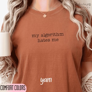 May include: A yam-colored t-shirt with the text "my algorithm hates me" in black. The word "yam" is printed below the text. The words "COMFORT COLORS" are printed in a pink box in the lower left corner.