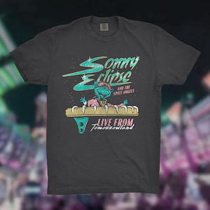 May include: Dark gray t-shirt featuring a retro design with the text "Sonny Eclipse and the Space Angels" and "Live From Tomorrowland." The graphic includes a cartoon alien character in a pink jacket and a teal hat.