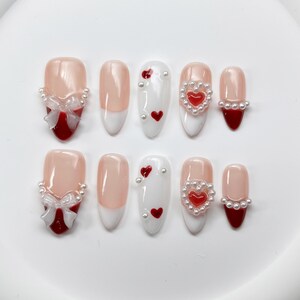 May include: Set of ten false nails with a Valentine's Day theme. The nails are decorated with red hearts, white pearls, and red bows.