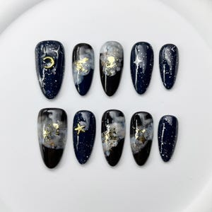 May include: Set of ten artificial nails with a dark blue base, featuring designs of gold foil, white clouds, and celestial accents like stars and a crescent moon. The nails have an oval shape and a glossy finish.
