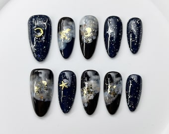 Op de afbeelding: Set van tien kunstnagels met een donkerblauwe basis, versierd met goudfolie, witte wolkenpatronen en hemelse motieven zoals sterren en een maan. De nagels zijn ovaal en hebben een glanzende afwerking.